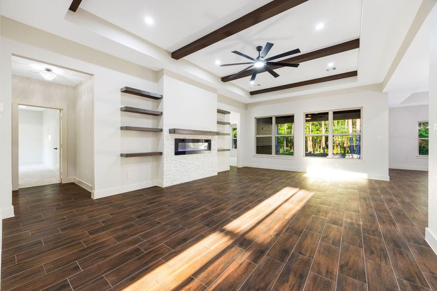 This spacious living area features elegant wood-look tile flooring, a modern electric fireplace, and built-in shelving. It boasts large windows for ample natural light and a ceiling fan with dark wood beams, adding a touch of sophistication.