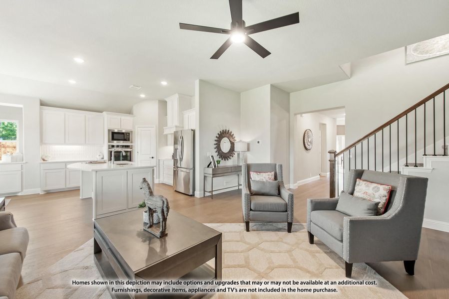 Open-concept living room with ceiling fan, hardwood floors, and white kitchen visible in background near staircase