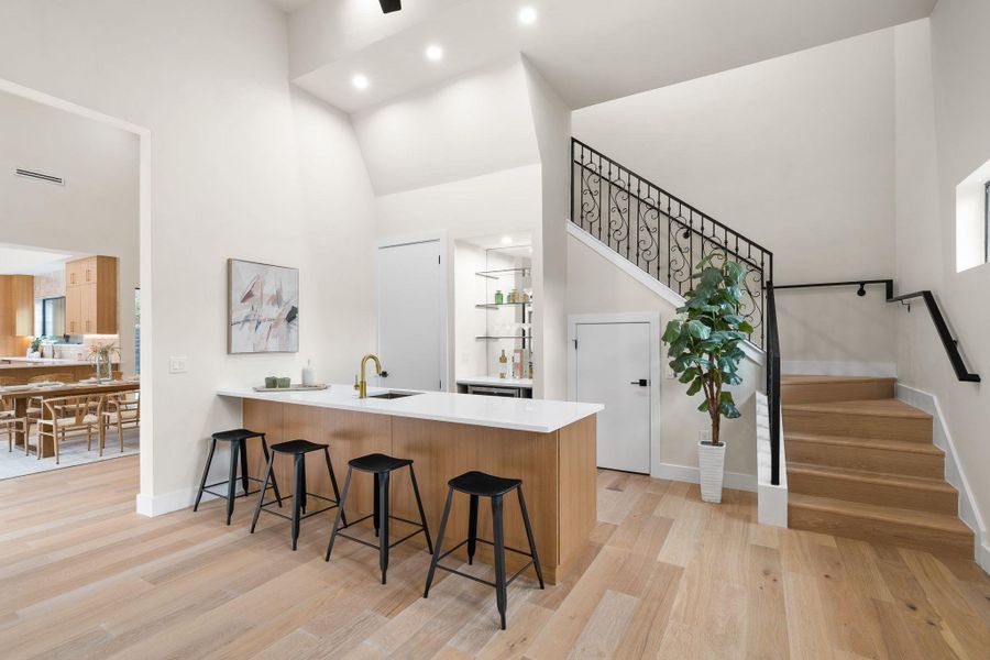 Modern interior featuring light wood-finish flooring, a built-in wet bar with a white countertop and gold-finish faucet, recessed lighting, and a grand staircase with wrought iron balusters