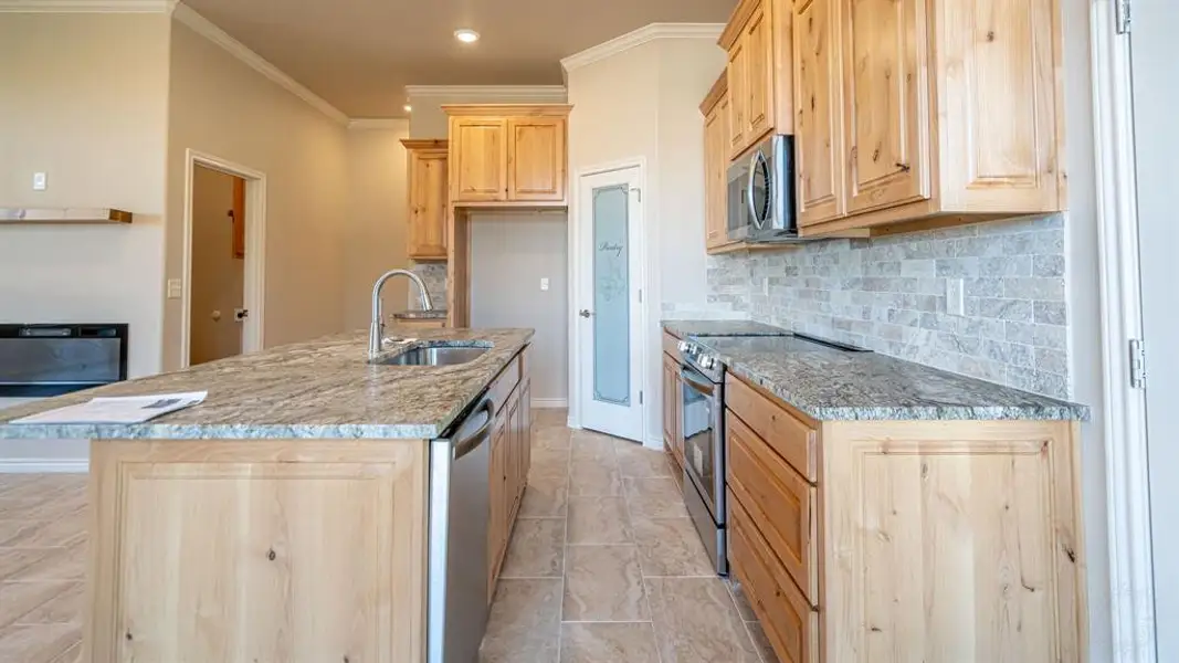 Kitchen featuring light brown cabinets, appliances with stainless steel finishes, ornamental molding, light stone counters, and an island with sink