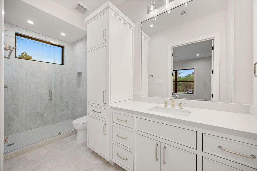 A bright and elegant bathroom designed with timeless finishes and thoughtful functionality, featuring a spacious vanity, abundant cabinetry, and sleek hardware. Natural light filters in, enhancing the clean, airy feel throughout. The highlight of this space is the oversized glass-enclosed walk-in shower with floor-to-ceiling tile and a built-in niche, creating a spa-like retreat perfect for everyday relaxation and comfort.