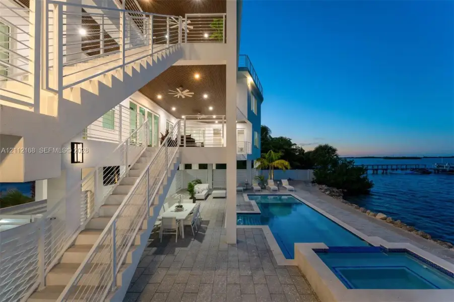Multi-level exterior featuring white cable-railing staircases, a covered patio with recessed lighting, an in-ground swimming pool, and an attached spa, situated directly on the waterfront Multi-level exterior featuring white cable-railing staircases, a covered patio with recessed lighting, an in-ground swimming pool, and an attached spa, situated directly on the waterfront