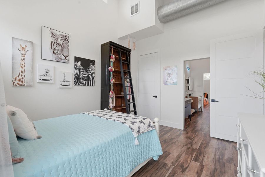 This bedroom features a cozy twin bed with a light blue quilt, hardwood floors, and animal-themed wall art. It includes a tall bookshelf with a ladder, bright white walls, and a doorway leading to another room. The space feels modern and inviting.