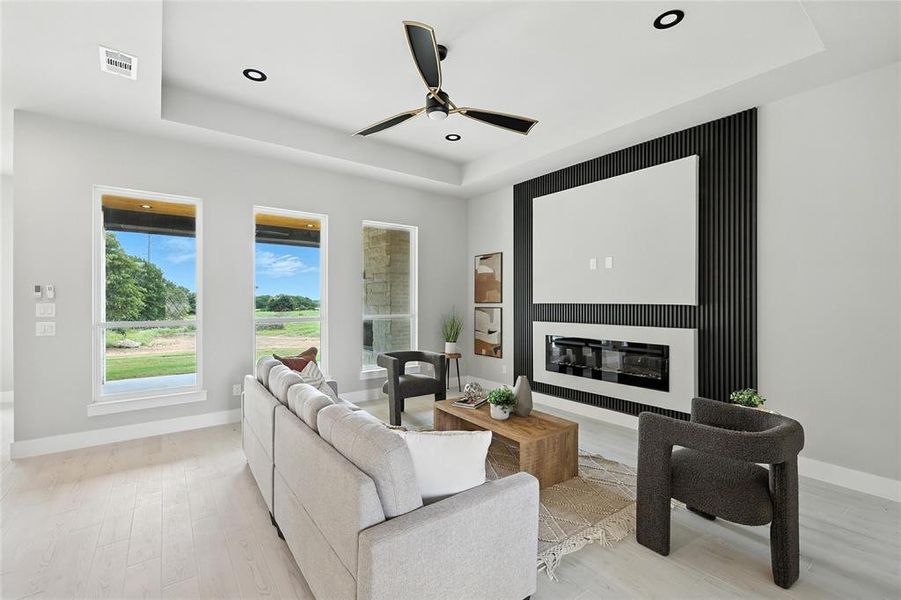 Living area featuring a raised ceiling, light wood-type flooring, ceiling fan, a glass covered fireplace, and recessed lighting