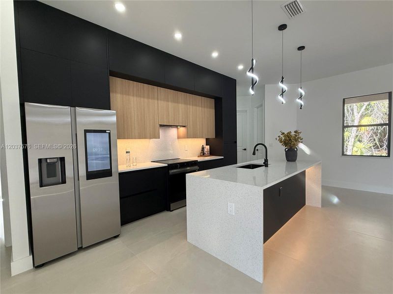 Contemporary kitchen featuring a large waterfall island with a black sink and matte black faucet, dark flat-panel cabinetry, light wood-finish upper cabinetry, and a stainless steel smart refrigerator