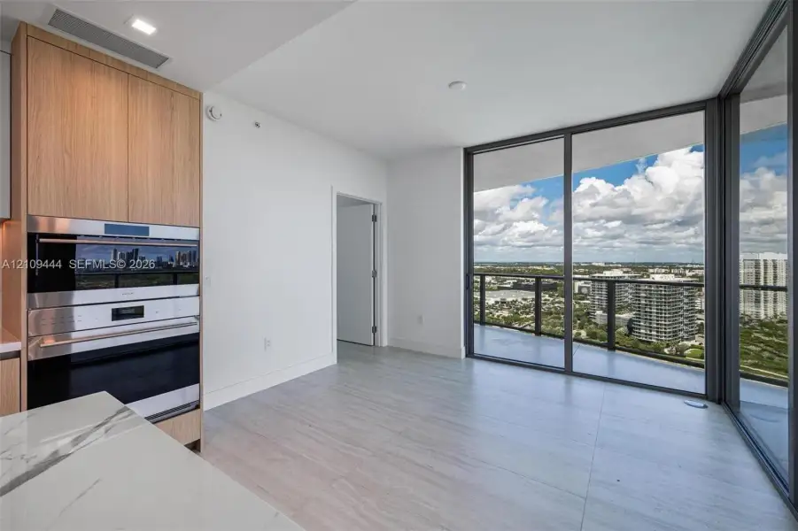 Floor-to-ceiling glass sliding doors, light-toned tile flooring, built-in wall oven and microwave, custom wood-finish cabinetry, and stone-surface countertop