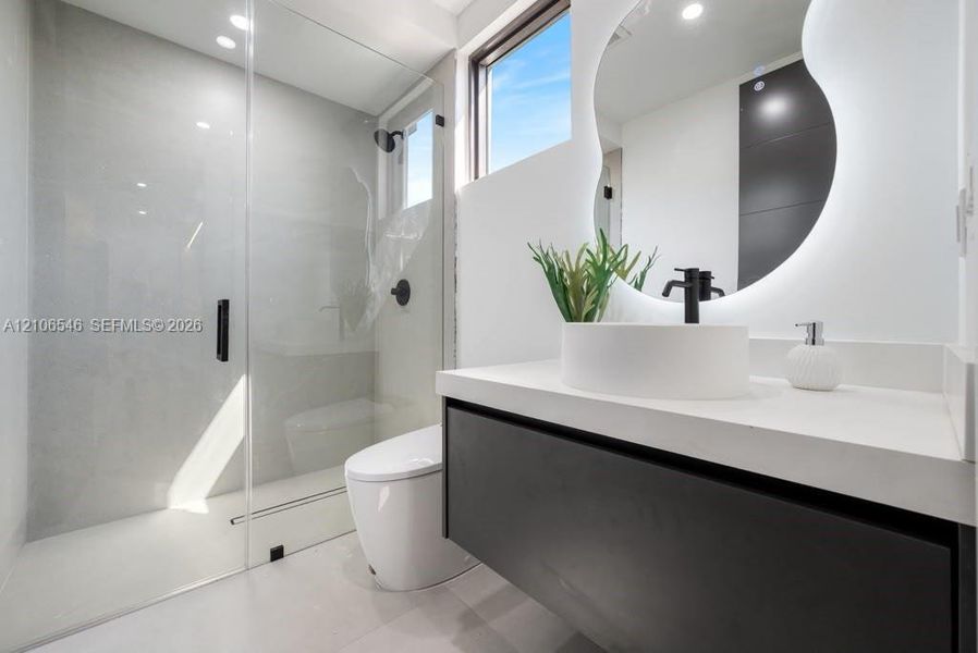 Floating dark-finish vanity with a white countertop and vessel sink, frameless glass walk-in shower with matte black fixtures, and a wall-mounted back-lit organic-shaped mirror