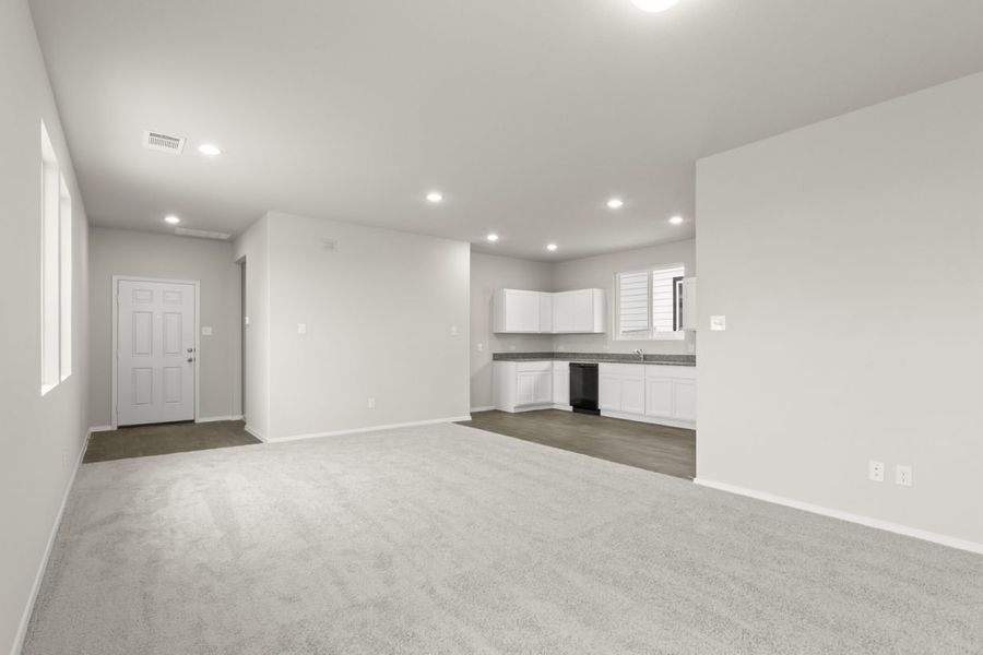 Image of a living room with cream walls, light grey carpeting, and a kitchen to the right with white cabinets
