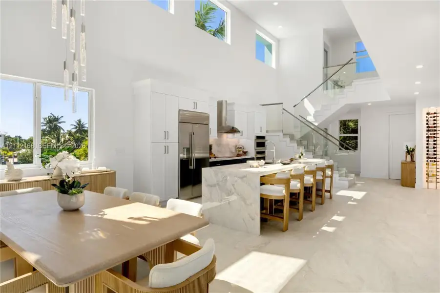 Open-concept kitchen featuring an expansive stone-topped waterfall island, custom white cabinetry, stainless steel professional-grade appliances, floating staircase with glass railings, and double-height ceilings with transom windows Open-concept kitchen featuring an expansive stone-topped waterfall island, custom white cabinetry, stainless steel professional-grade appliances, floating staircase with glass railings, and double-height ceilings with transom windows