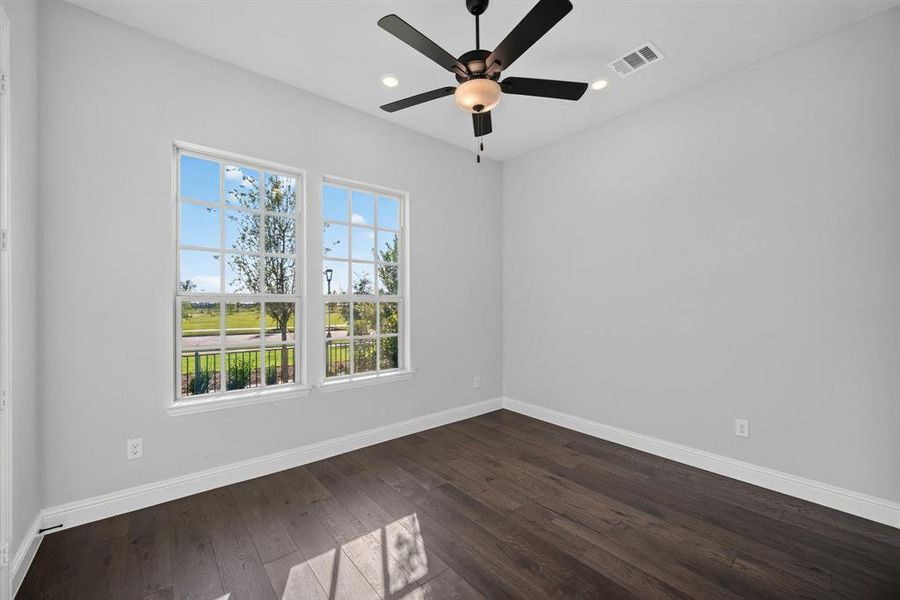First floor bedroom/study featuring two large windows, beautiful wood-finish flooring, light gray walls, closet and a ceiling fan with integrated lighting