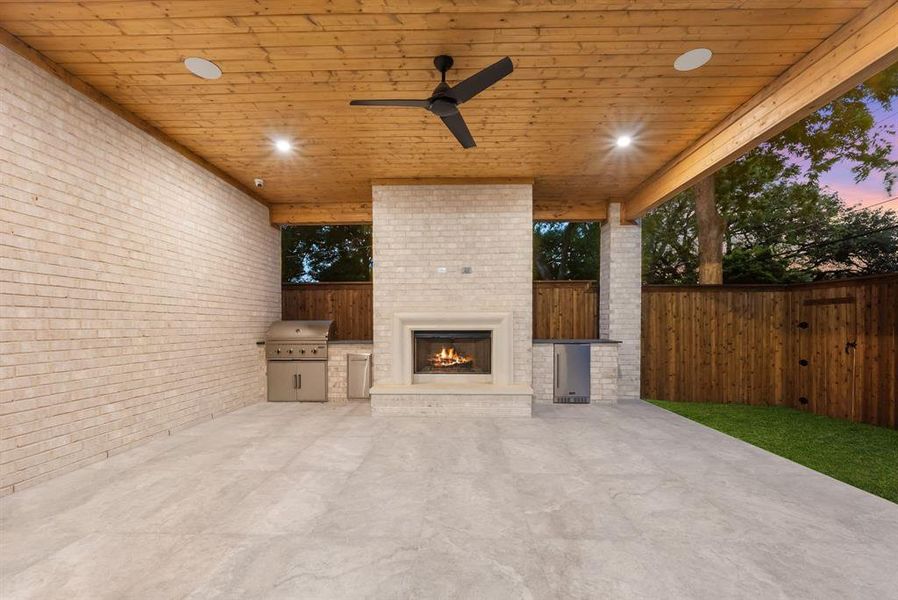 This outdoor living space features a wooden plank ceiling with recessed lighting and a ceiling fan, a built-in fireplace, a stainless steel grill, and a stainless steel mini-fridge
