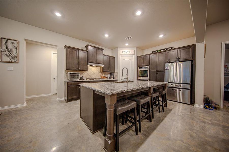 This modern kitchen features dark wood cabinetry, granite countertops, and stainless steel appliances. It includes a spacious island with bar seating, recessed lighting, and a pantry for ample storage.