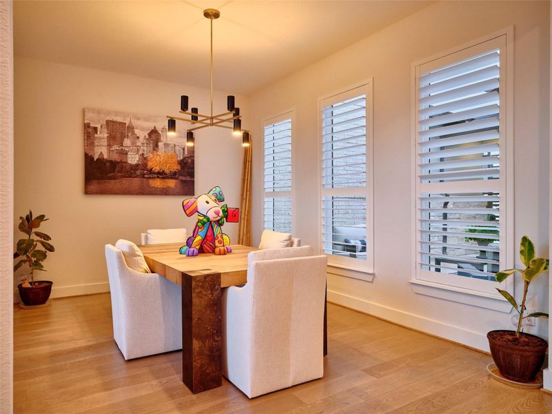 Elegant open dining area with a striking light fixture, warm flooring, and designer shutters overlooking a lavish courtyard.