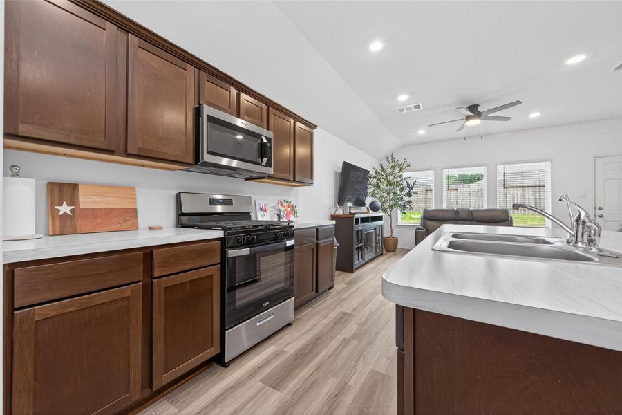 This modern open-concept kitchen and living area features sleek stainless steel appliances, dark cabinetry, and a large island with a sink. The space is bright and welcoming with large windows, recessed lighting, and a ceiling fan, creating a comfortable atmosphere for entertaining or relaxing.