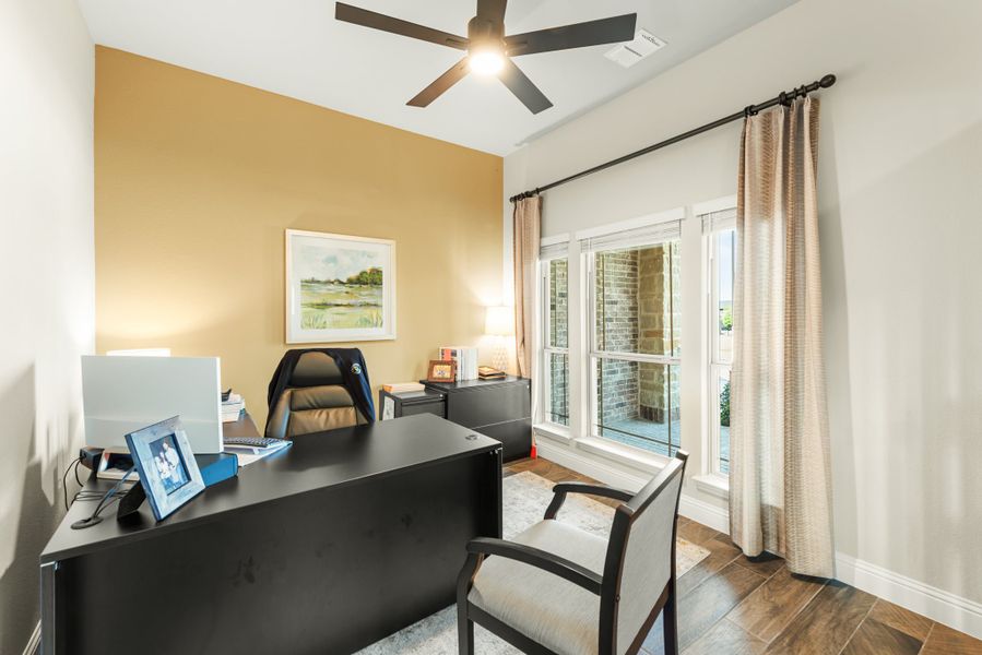 Home office with dark wood desk, ceiling fan, accent wall, and large window with curtains