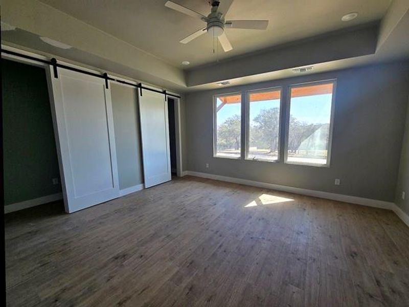 Unfurnished bedroom with a barn door, a tray ceiling, dark wood-style flooring, and a ceiling fan