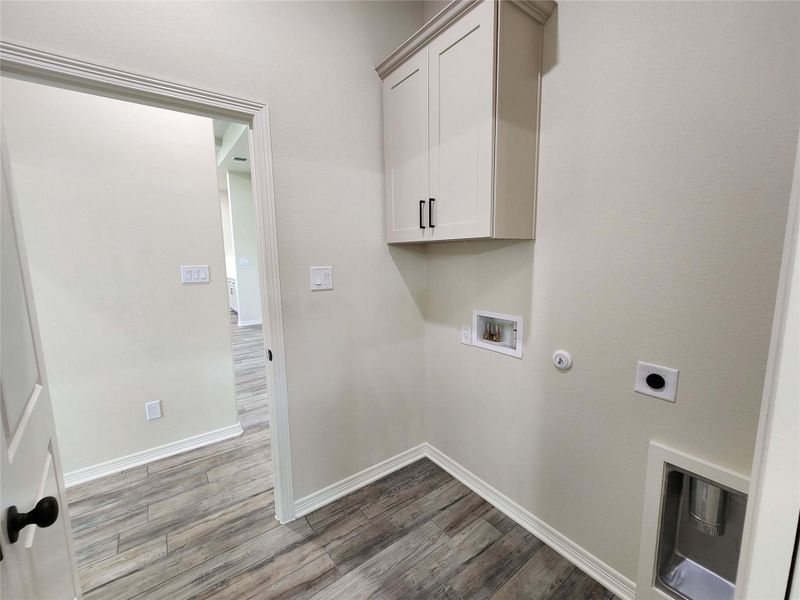 Dedicated laundry area featuring wood-finish flooring, light-toned walls, overhead cabinetry with dark hardware, and hookups for appliances
