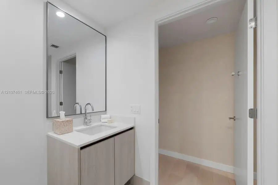 Bathroom vanity featuring light-toned cabinetry, a solid surface countertop with an integrated rectangular sink, a polished chrome bridge faucet, and a large rectangular mirror with a thin metallic frame Bathroom vanity featuring light-toned cabinetry, a solid surface countertop with an integrated rectangular sink, a polished chrome bridge faucet, and a large rectangular mirror with a thin metallic frame