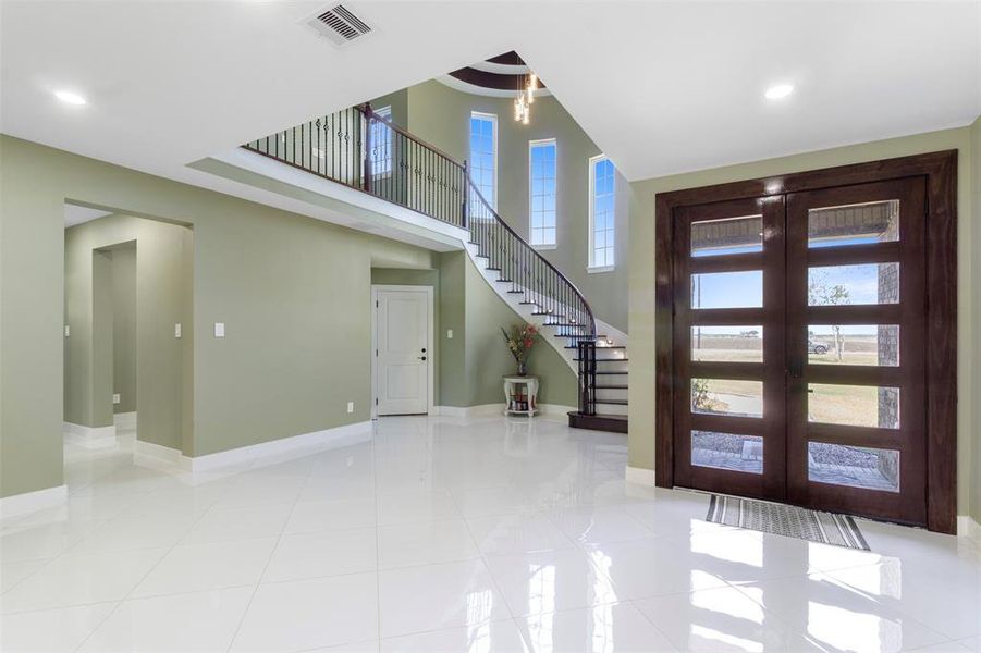 This spacious entryway features high ceilings and abundant natural light from tall windows. It includes a striking curved staircase with elegant railings and glossy white tile flooring. The large wooden front door adds a touch of sophistication, complementing the room's open, airy feel.