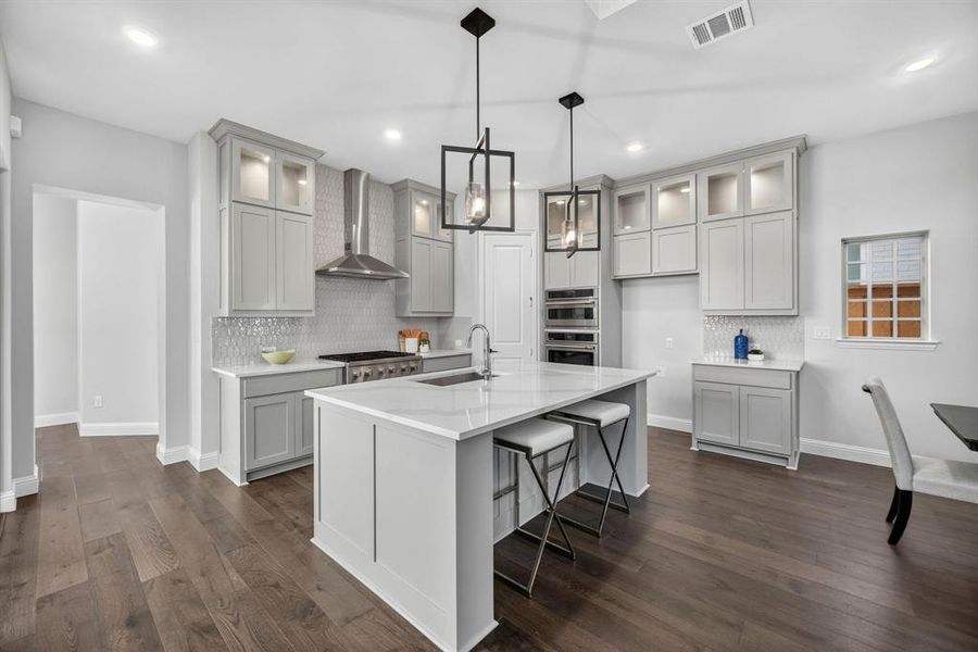 Modern kitchen featuring a large central island with seating, wood-finish flooring, light gray cabinetry with glass accents, and a white tile backsplash