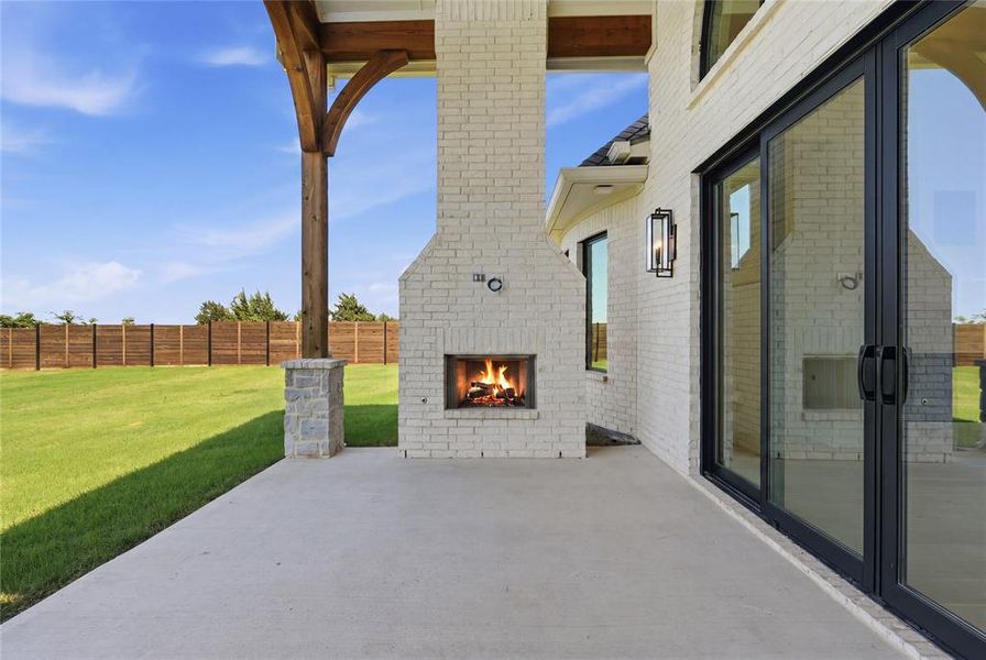 Expansive concrete patio featuring a white brick outdoor fireplace, black-framed sliding glass doors, and a wood beam supported by a stone column