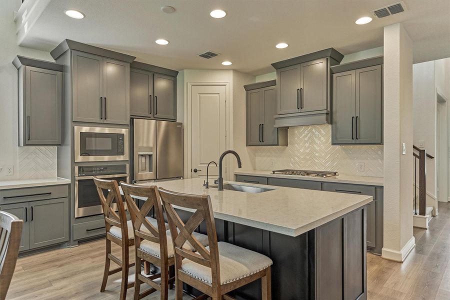 Modern kitchen with sleek gray cabinets, stainless steel appliances, and a central island with seating. Bright lighting and herringbone backsplash enhance the contemporary design.