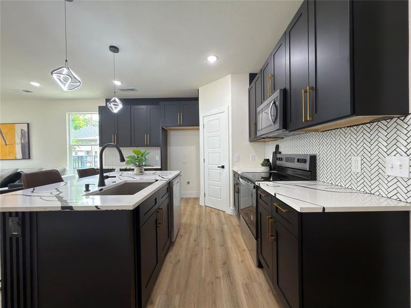 Contemporary kitchen featuring dark cabinetry with gold-tone hardware, white countertops, a herringbone tile backsplash, stainless steel appliances, and wood-finish flooring