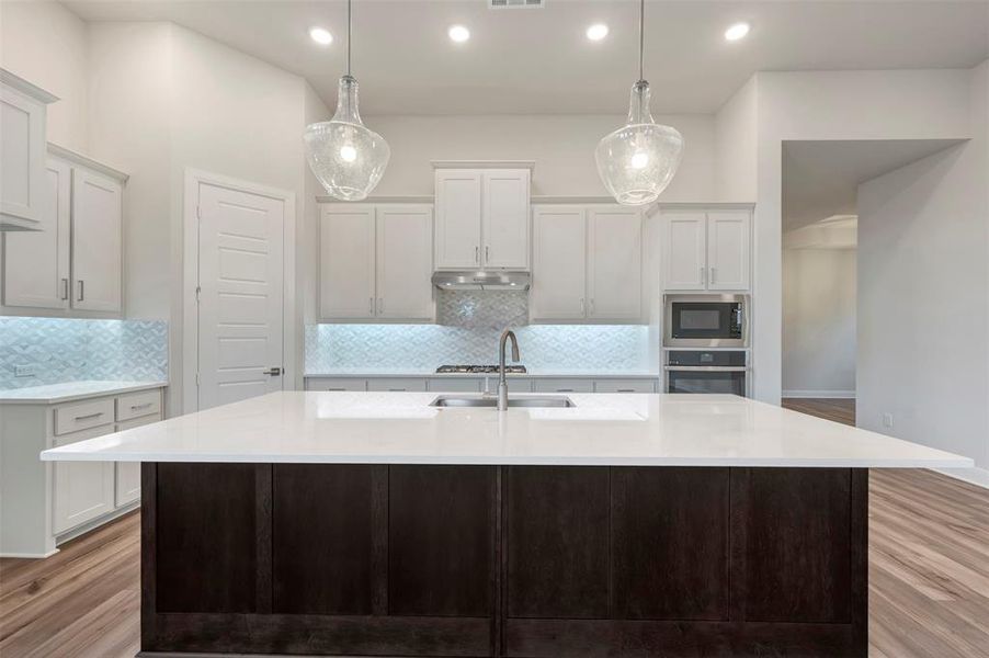 Two tone kitchen featuring light wood-type flooring, tasteful backsplash, light stone countertops, an island with sink, and dual tone cabinets