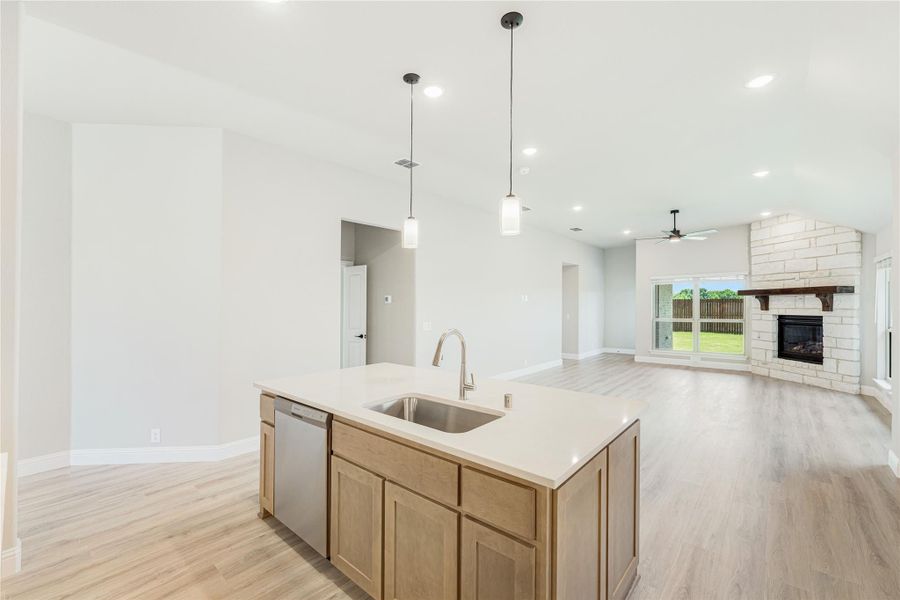 Kitchen island with sink and pendant lights open to living room with stone fireplace and light wood floors