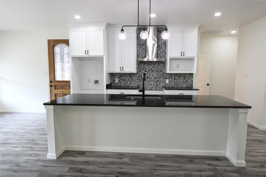 Kitchen featuring white cabinets, wall chimney range hood, a large island with sink, dark wood finished floors, and backsplash Kitchen featuring white cabinets, wall chimney range hood, a large island with sink, dark wood finished floors, and backsplash