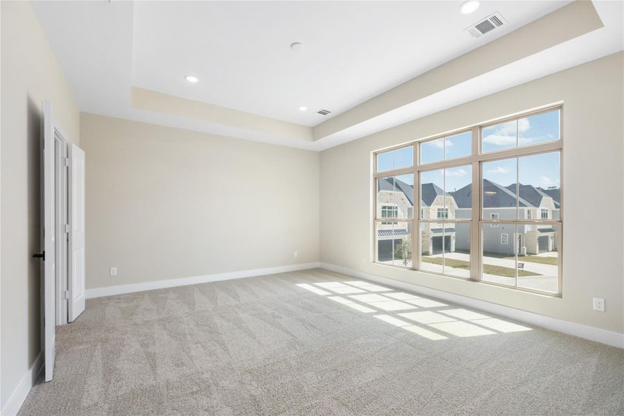 As you enter the spacious primary bedroom, you're greeted by a cozy and inviting atmosphere. Natural light floods the room through multiple windows, and a view into the adjacent space with large windows adds to the open feel. The elegant tray ceiling enhances the room's character, making it the perfect retreat, whether for rest, relaxation, or personalization.