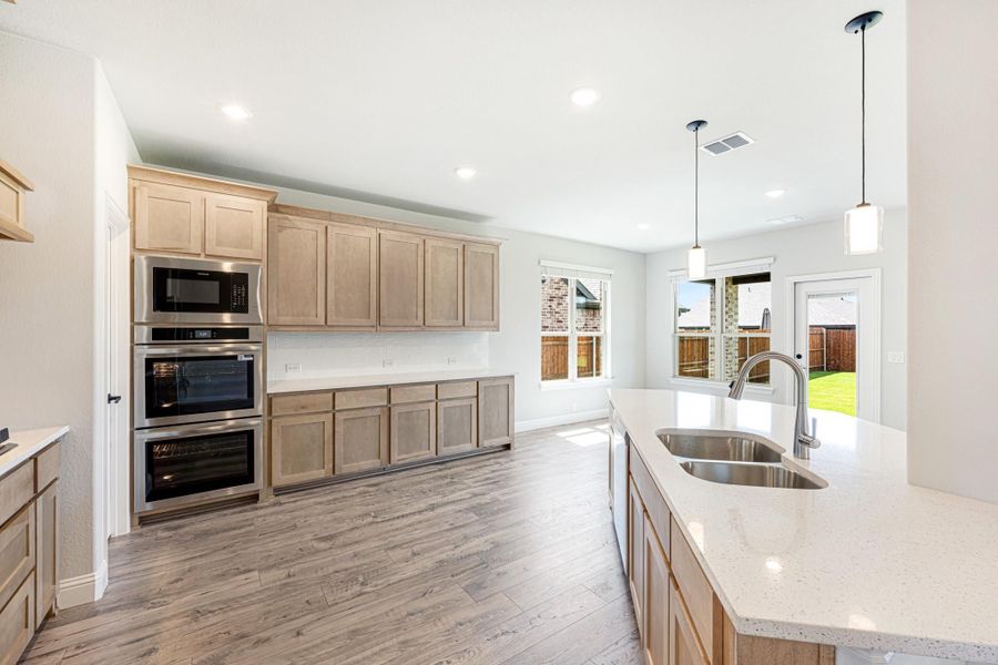 Kitchen with light wood cabinets, double wall oven, quartz island with sink, and pendant lights over hardwood floors
