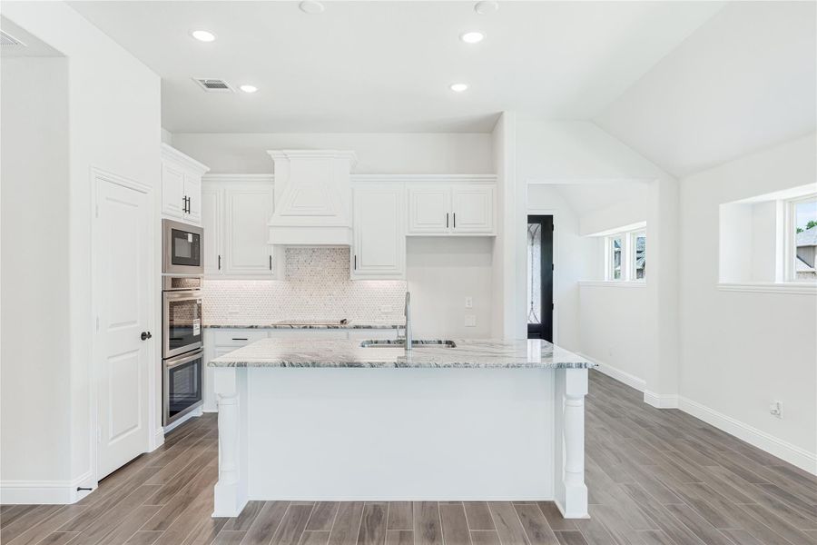 White kitchen with large island, granite countertops, stainless steel ovens, and tile backsplash on wood-look floors White kitchen with large island, granite countertops, stainless steel ovens, and tile backsplash on wood-look floors