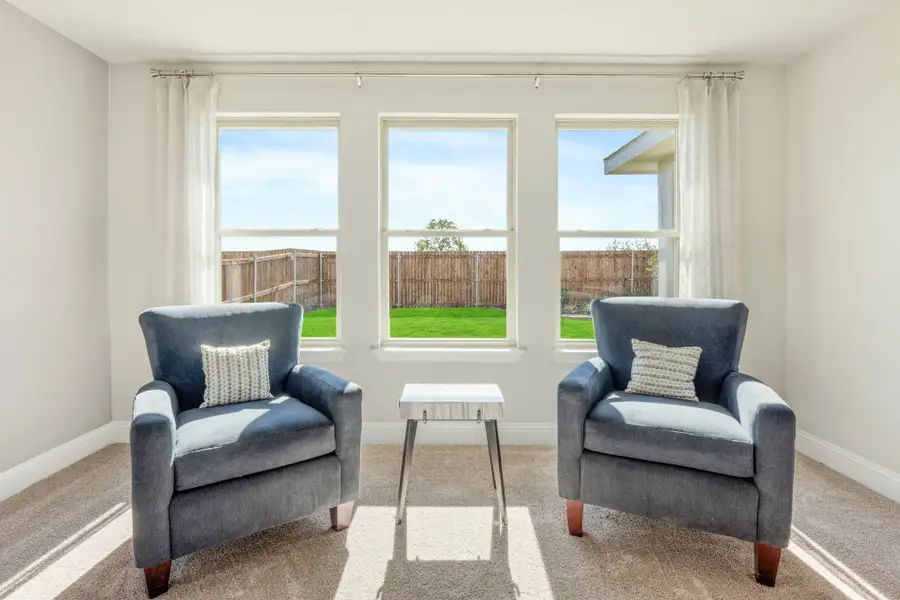 Sitting area with two blue armchairs, small side table, and three windows overlooking a fenced backyard