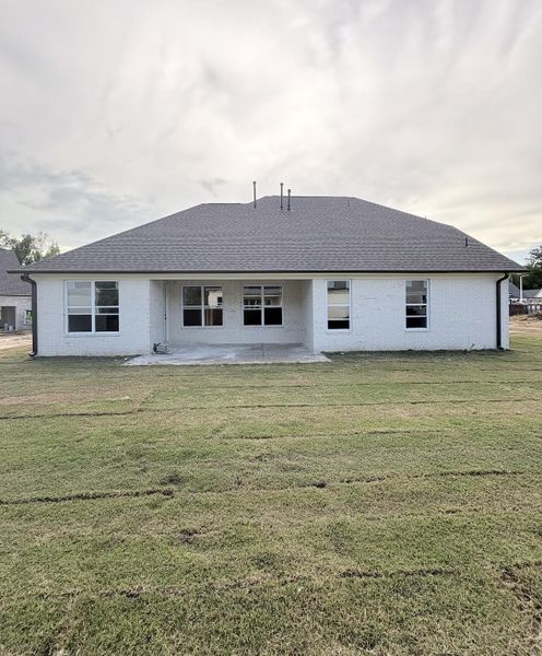 White brick exterior with a dark shingle roof, a covered concrete patio, multiple windows, and a green lawn
