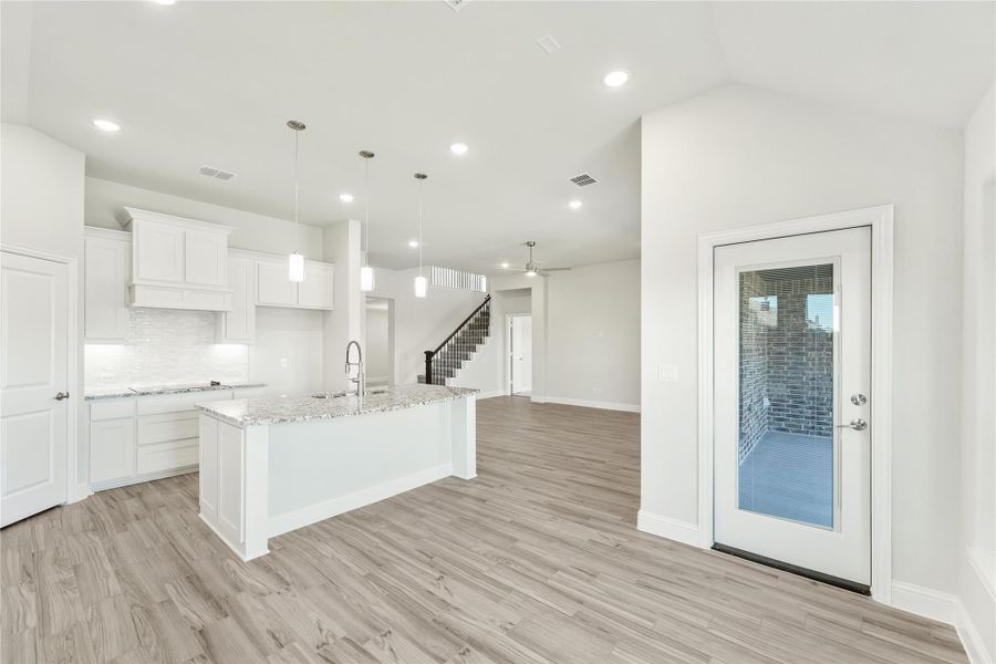 Open kitchen with white cabinets, granite island, and light wood floors leading to living area and staircase