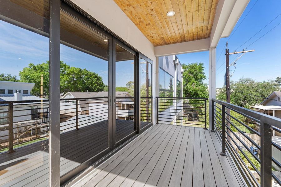 The covered second-floor balcony is finished with a tongue-and-groove wood ceiling and recessed lighting — the same material warmth found in the porte-cochere below. Large sliding glass doors connect directly to the interior and black cable railings keep the treetop views fully open.