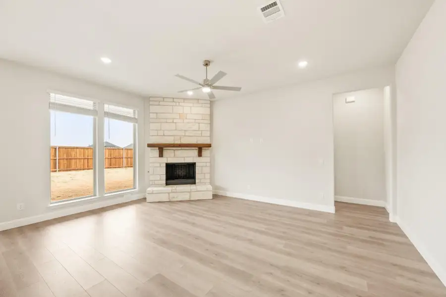 Open living room with stone corner fireplace, wood mantel, ceiling fan, and light hardwood floors Open living room with stone corner fireplace, wood mantel, ceiling fan, and light hardwood floors