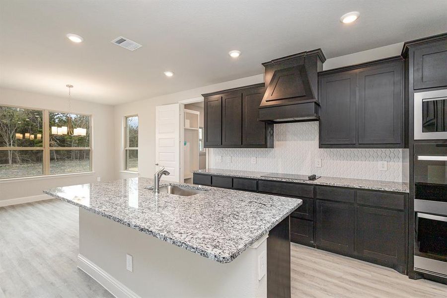 Kitchen with black electric cooktop, premium range hood, a center island with sink, and sink