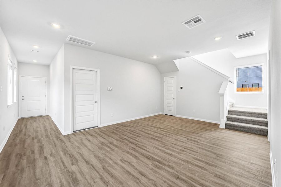 Bright and spacious living area with modern wood-look flooring, recessed lighting, and a neutral color palette. Features include a staircase with carpeted steps, multiple doors, and ample natural light from a window. Please note: The image is not the actual home; it is a home with the same floor plan in the neighborhood. Finishes may vary. Bright and spacious living area with modern wood-look flooring, recessed lighting, and a neutral color palette. Features include a staircase with carpeted steps, multiple doors, and ample natural light from a window. Please note: The image is not the actual home; it is a home with the same floor plan in the neighborhood. Finishes may vary.