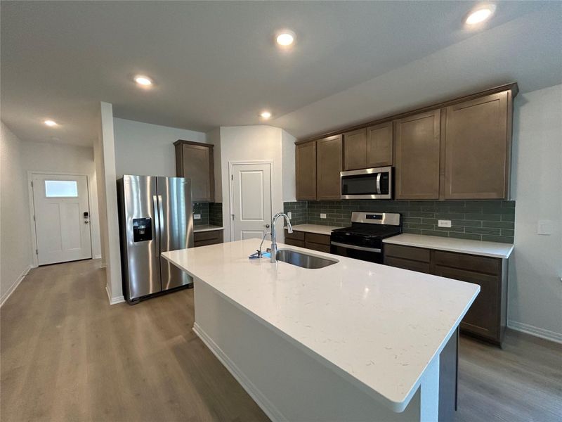 Kitchen featuring appliances with stainless steel finishes, backsplash, light stone counters, an island with sink, and dark wood-style floors