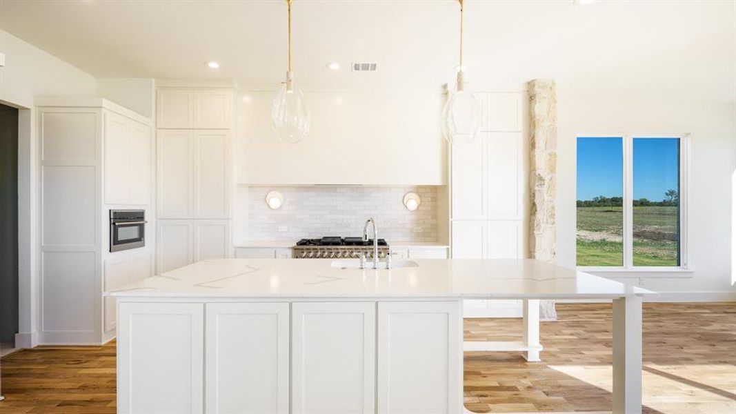 Kitchen with light stone countertops, sink, pendant lighting, light hardwood / wood-style flooring, and a kitchen island with sink