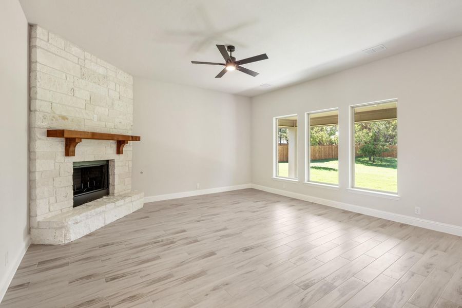 Empty living room with stone corner fireplace, wood mantel, ceiling fan, and large windows overlooking backyard