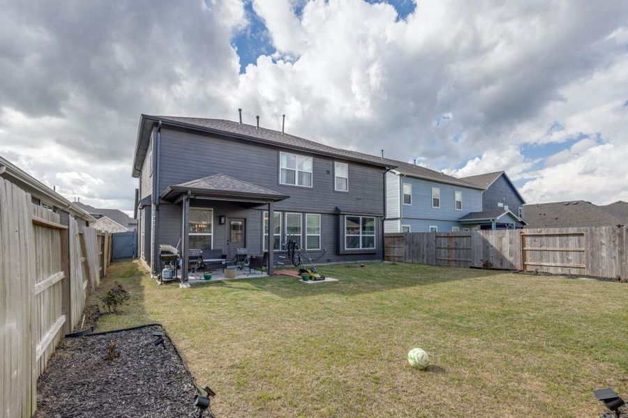 A spacious backyard with a well-maintained lawn, bordered by a wooden fence. The two-story home features a covered patio area, perfect for outdoor seating. The exterior is modern with large windows, enhancing natural light inside.