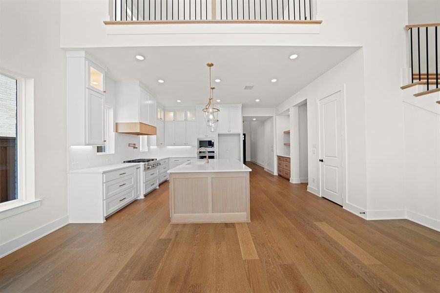 Kitchen featuring an island with sink, light wood-type flooring, glass insert cabinets, decorative light fixtures, and white cabinetry Kitchen featuring an island with sink, light wood-type flooring, glass insert cabinets, decorative light fixtures, and white cabinetry