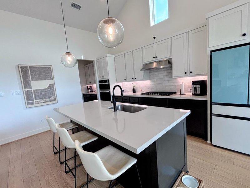 Modern kitchen features sleek white cabinetry, a spacious island with a sink, and stylish pendant lighting. It includes stainless steel appliances, a subway tile backsplash, and ample natural light, creating a bright and inviting space.