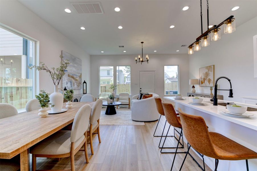 View from dining room through to living area and front entry, showing the full open-concept flow, hardwood floors, recessed lighting, island bar seating with leather stools, and chandelier over dining table.