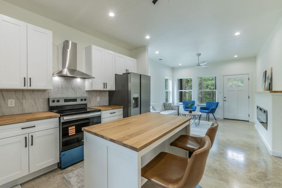 Open-concept kitchen featuring white shaker cabinetry, butcher block countertops, stainless steel appliances, a center island with seating, and concrete flooring Open-concept kitchen featuring white shaker cabinetry, butcher block countertops, stainless steel appliances, a center island with seating, and concrete flooring