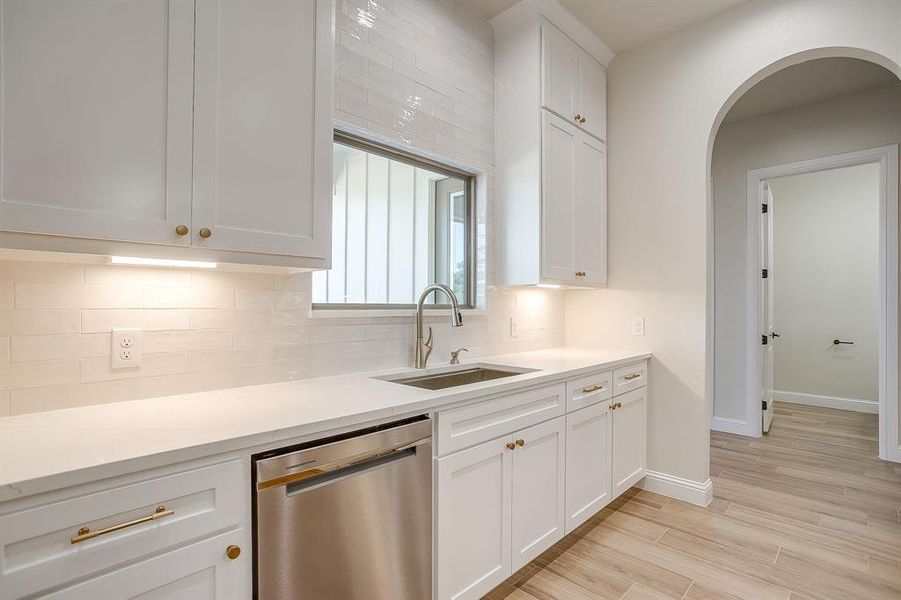 Kitchen featuring stainless steel dishwasher, tasteful backsplash, white cabinets, light wood-type flooring, and light stone countertops Kitchen featuring stainless steel dishwasher, tasteful backsplash, white cabinets, light wood-type flooring, and light stone countertops