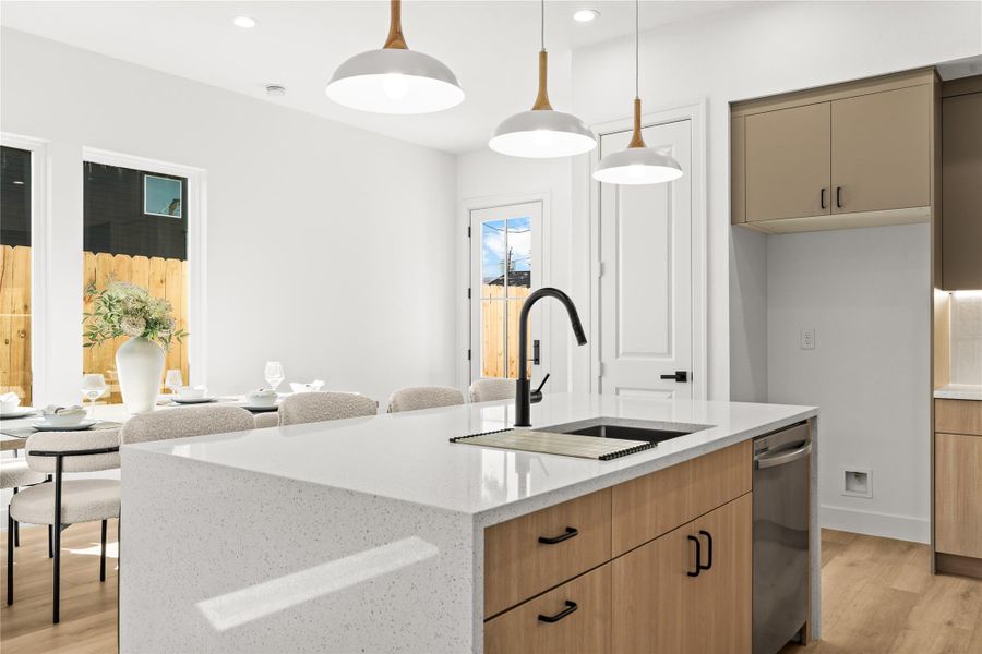 The kitchen island features a white quartz countertop with a deep, under-mount stainless steel sink and a matte black pull-down faucet. A stainless steel dishwasher is built right into the natural wood lower cabinets directly across from the sink, making cleanup easy.